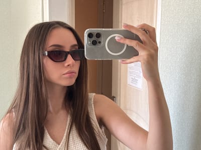A young woman with long brown hair is taking a selfie with a phone in a light-colored room. She is wearing dark sunglasses and a white top. The background consists of a door and patterned wall. The location appears to be indoors, possibly a room in Береговой сельский совет, Ukraine, although the exact address cannot be determined from the image.