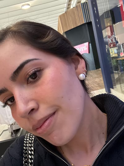 A close-up, angled selfie of a woman with brown hair and defined eyebrows. She has dark eyes with long eyelashes. Her skin tone appears fair with some visible freckles. She is wearing a pearl earring with a sparkling band. She is also wearing a dark, possibly corduroy jacket with a zipper and chain detail. The background shows a blurred reflection in a window of what appears to be a store or commercial building. The location is not provided in the original description, and it is not possible to determine the location in the city of San Francisco Ocotlán (Ocotlán), Mexico from the image alone.