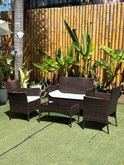 The scene depicts an outdoor patio area in Panglao, Philippines, illuminated by direct sunlight. The ground is covered with green artificial turf, marked by a dark linear seam running horizontally across the midground. A four-piece dark brown woven rattan furniture set is arranged on the turf, comprising a two-seater sofa and two single armchairs, each fitted with a white cushion, and a rectangular coffee table with a dark, reflective top surface. Behind the furniture, a tall, vertical bamboo fence forms the primary background, fronted by several large-leafed tropical plants resembling banana trees, interspersed with smaller variegated foliage and potted plants. To the far left, a palm tree trunk is visible, with a white patio umbrella partially seen behind it and a white, circular, wall-mounted object attached to the tree trunk. Shadows from the furniture are distinctly cast onto the artificial turf. No individuals are present in the image.