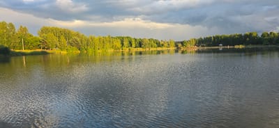 This is a wide, panoramic, eye-level photograph of a serene lake and its tree-lined shore. The sun is low, casting a warm golden light on the lush green trees and the rippling surface of the water, creating a tranquil and picturesque scene. The sky is a mix of dramatic, dark storm clouds and patches of brighter, illuminated sky, suggesting an approaching or receding weather front. On the far shore, a line of trees forms a dense natural barrier, with hints of a wooden fence and a small white gazebo visible amongst the foliage in the distance, suggesting human presence or a recreational area. The water itself is calm, with gentle waves reflecting the surrounding environment and the sky above. The photograph is taken from what appears to be a dock or the edge of the water, with a slight blur in the foreground suggesting the edge of an object. The scene conveys a sense of natural beauty and peacefulness, characteristic of a rural or park-like setting. The location is identified as the city of Dubrovka, Russia.