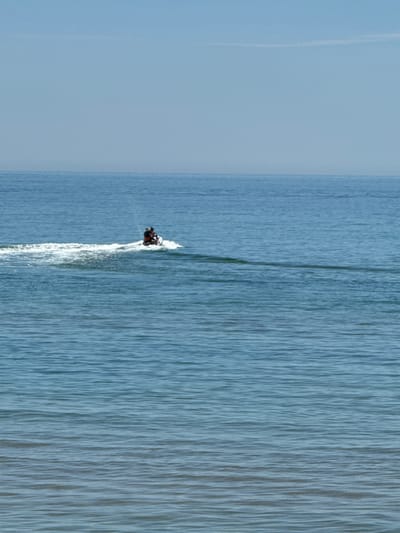 The image, when viewed in its correct orientation, captures a serene daytime scene of a person engaged in water sports. The horizon line bisects the frame horizontally, separating a clear sky above from an expansive body of water below.

The primary subject is a single individual riding a personal watercraft, likely a jet ski, in the mid-right portion of the image. The jet ski appears dark-colored, possibly black and red, and is actively moving, leaving a distinct white, foamy wake that stretches diagonally behind it towards the lower-left. The rider, dressed in dark clothing, is seated on the craft, facing towards the right side of the frame.

The water surface is a calming blue-grey, textured by countless fine, parallel ripples or small waves that extend across the entire visible expanse, suggesting gentle movement. The sky above is a uniform, pale blue, devoid of clouds, indicating bright and clear weather conditions. A faint, almost transparent line, possibly a distant contrail, is barely visible in the upper-left quadrant of the sky.

The scene portrays a moment of recreational activity on open water, consistent with a coastal setting such as Frankston, Australia. The time of day appears to be daytime, under sunny and favorable weather. No specific emotions are discernible from the distant rider, and no text is visible within the image.