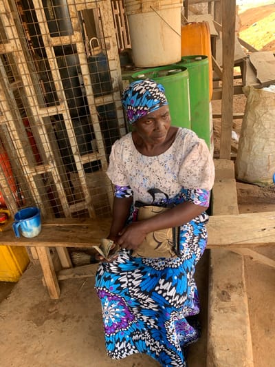 An adult female is seated on a light-colored wooden bench in a semi-enclosed outdoor area within Ube, Nigeria. She wears a vibrant blue, purple, white, and black patterned headwrap and a matching long skirt, with a white, lace-textured, short-sleeved top over an undergarment featuring the same pattern visible at her forearms. Her left hand holds a fanned stack of banknotes. A light brown, patterned shoulder bag rests on her lap. To her immediate left on the bench is a blue and white plastic mug. Behind her, a rusty metal grate structure with a padlock is positioned to the left, while several green plastic drums, an orange plastic jerrican, and a white plastic bucket are stacked to her right. A large, light-colored sack is visible further to the right. The ground surface is uneven earth. The woman's gaze is directed towards the right side of the frame.
