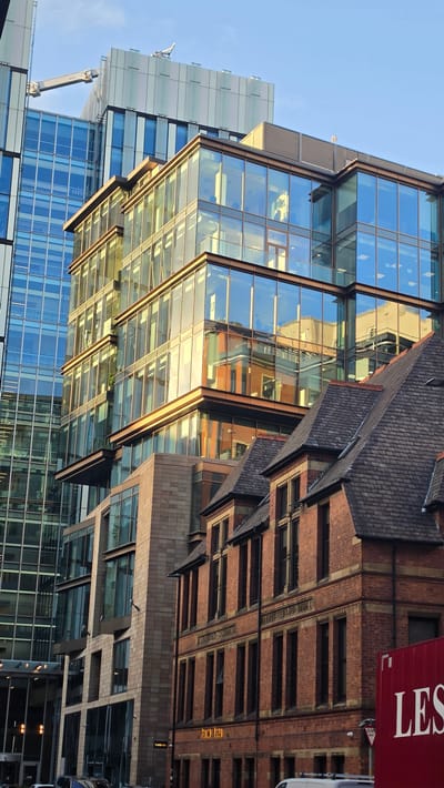 This is a photograph of a street scene in Manchester, United Kingdom, featuring a juxtaposition of modern glass buildings and a traditional brick building. The modern structures on the left are tall, with extensive glass facades that reflect the sky and surrounding environment. One building has a distinctive cantilevered section with gold-colored trim. In the foreground, on the right, is an older brick building with a pitched, dark-tiled roof and numerous dark-framed windows. Inscriptions are visible on this brick building, including "FRIENDS SCHOOL" and "ERECTED AD 1889". A large red container with white lettering, "LES", is partially visible on the far right. The sky is blue with some wispy clouds. The lighting suggests it is daytime, possibly late afternoon, with the sunlight casting warm tones on the modern building's glass and the brickwork. There are no discernible people or vehicles in immediate focus, though the lower portions of buildings suggest a street level environment with potential for traffic.