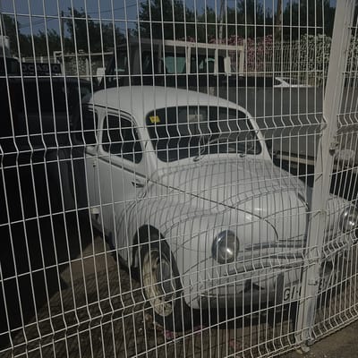 The image captures a white vintage car, possibly a Renault 4CV, parked behind a wire fence. The car is predominantly visible, with its rounded hood, headlights, and front grille partially obscured by the fence. A yellow sticker is affixed to the windshield. The fence itself is a light gray metal mesh with vertical supports. Beyond the car and fence, trees and some pink and white flowering bushes are visible. In the background, a white vehicle, possibly a van, is also present. Part of a sign reading "OCASIO" is visible on the left side of the frame, suggesting a used car lot or similar establishment. The ground appears to be dirt or gravel, with some debris scattered around the base of the car. The lighting suggests it is daytime, with shadows cast by the fence onto the car and the ground. The overall setting appears to be an outdoor storage or parking area, potentially in the city of El Perelló, Spain, given the context. There are no people visible in the image.