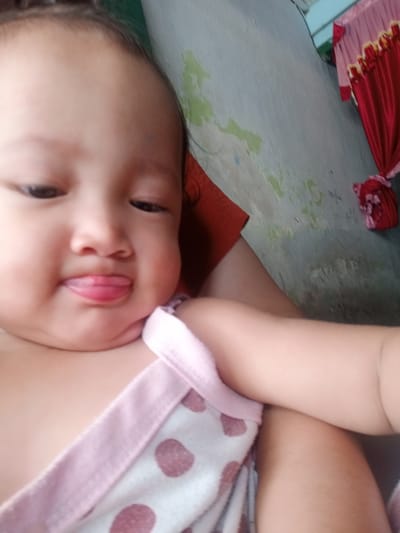 A baby is being held in someone's arm. The baby is wearing a shirt with pink polka dots. The baby is sticking out their tongue. Behind the baby and arm, the wall is white and gray, and a red curtain hangs in the background. The location is Bungur, Indonesia.