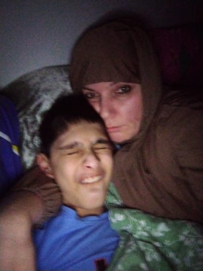 The image shows a close-up selfie of two individuals, an adult female and a child, lying down in what appears to be a domestic setting. The female, on the right, is wearing a brown head covering and a brown garment, possibly a hooded sweatshirt. She is looking towards the camera with her left eye visible. The child, on the left, is a young boy with dark hair, wearing a blue t-shirt. His eyes are closed and his mouth is open in an expression that could be laughter or discomfort. They are both partially covered by a green and white patterned blanket. The lighting is dim, suggesting it might be nighttime or a dimly lit room. The background is dark and indistinct, with some texture visible that could be bedding or furniture. There are no discernible landmarks or elements that would place this scene specifically within Al Mas'oudi, United Arab Emirates. The image captures an intimate moment between the two individuals.