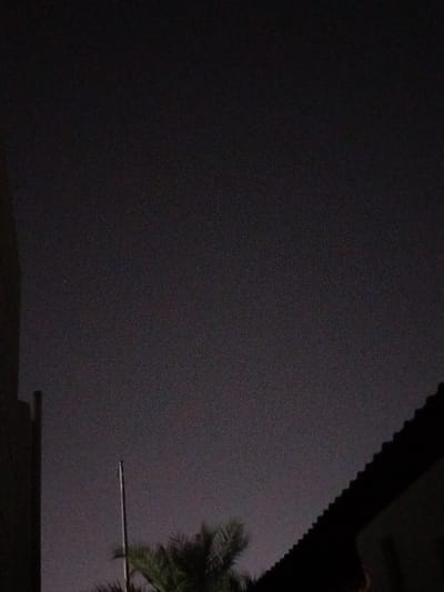 This is a nighttime photograph taken in Al Mas'oudi, United Arab Emirates. The image is largely dark, with a textured, grainy appearance. The sky is a dark, overcast gray. In the foreground, the dark silhouette of a roofline is visible on the right side of the frame, angled upwards. To the left of the roofline, a tall, thin, light-colored pole or antenna extends vertically into the sky. Below and to the left of the pole, the dark green fronds of a palm tree are visible. On the far left of the image, a dark vertical structure, possibly a building or wall, is partially visible. The scene appears to be an outdoor urban or suburban setting at night.