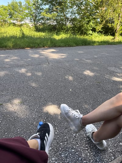 The image shows a section of asphalt road in Elhovo, Bulgaria, bordered by a grassy area with trees. Three pairs of legs and shoes are visible: one pair with white sneakers, one with white platform sneakers, and another with black Adidas sneakers and a water bottle next to the right foot. The perspective is directly above the legs, looking downward onto the road. Sunlight is present, casting shadows.
