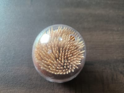 A clear, spherical plastic container filled with sharpened wooden toothpicks sits on a dark wooden surface. The container is positioned slightly off-center, towards the right side of the frame. The toothpicks are densely packed and point upwards, creating a textured, spiky surface within the sphere. Light reflects off the curved surface of the container, obscuring some of the toothpicks. The background is a dark, textured wood grain, appearing to be a table or countertop. The overall lighting suggests an indoor environment. No people or animals are present in the image.