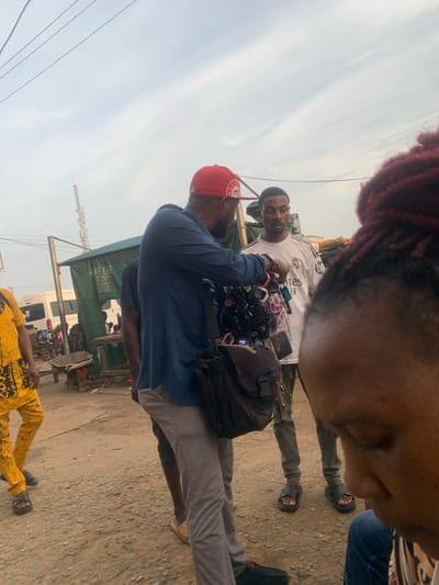The image shows a street scene in Elelenwo, Nigeria, with several individuals interacting in what appears to be a market environment. In the foreground, a person's head and shoulder are visible, facing away from the camera. To the left, a person wearing a yellow and black patterned outfit and black sandals is walking away. In the center, a man in a navy blue shirt, gray pants, and a red cap is showing a display of sunglasses to another man who is wearing a white Real Madrid jersey, gray pants, and black sandals. The man in the jersey is looking at the sunglasses. Behind them, a green tent or stall is set up, with text visible on it, and a white van is parked nearby. The ground is unpaved and appears to be dirt and gravel. A telecommunications tower is visible in the background against a cloudy sky. The overall scene suggests a bustling marketplace with vendors and customers.