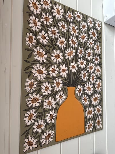 The image shows a close-up of a painting or mural featuring a bouquet of white daisies with brown and orange centers, arranged as if spilling out of a rounded, tan-colored vase. The artwork is set against a muted green background, with dark green leaves and thin, dark outlines accentuating the flowers and their stems. The style is graphic and somewhat stylized, with bold lines and flat areas of color. The painting appears to be leaning against a white, paneled surface, suggesting it might be displayed indoors or on a covered patio. No people are visible. The lighting is even, indicating it is likely daytime, but there are no specific weather cues. The overall mood is cheerful and decorative, emphasizing the floral motif. There is no visible text in the image.