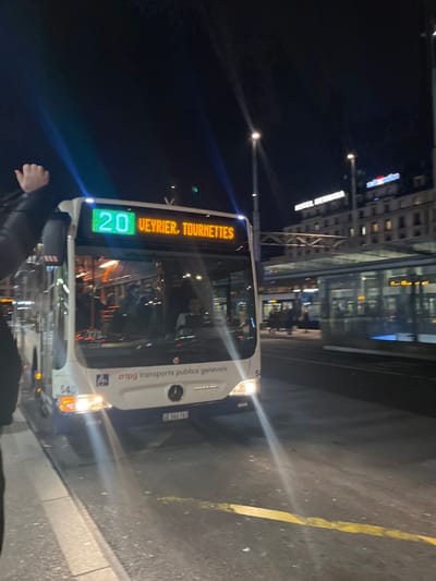 This night-time image, taken in Genève, Switzerland, shows a white city bus stopped at what appears to be a bus stop or street-side location. The bus, possibly a Mercedes-Benz (indicated by the logo), has its headlights on, illuminating the immediate area. Its electronic destination display clearly shows "20" in green for the route number and "VEYRIER, TOURNETTES" in amber for the destination.
The side of the bus features the text "tpg transports publics genevois", identifying it as part of Geneva's public transport, along with a wheelchair accessibility symbol. A unit number "543" is visible on the front, and the license plate "GE-900763" further confirms the location as Geneva.
In the foreground, the arm and shoulder of a person wearing a dark jacket are visible. The background includes several illuminated buildings, suggesting an urban environment. A distinctive red illuminated sign, likely "Swisscom", is visible on one building, along with other less discernible white illuminated signs. Streetlights punctuate the dark sky, casting rays of light. The ground is dark asphalt with a yellow painted line. The scene depicts a quiet moment in a city at night.