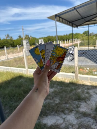 A light-skinned human hand, with pink manicured fingernails, holds four fanned-out tarot cards against an outdoor backdrop. A small black tattoo of two upward-pointing chevron symbols is visible on the inner wrist. The prominent rightmost card is "The Sun" from a Rider-Waite-style deck, depicting a large yellow sun, a naked child on a white horse, and sunflowers. The other three cards, partially obscured, also feature celestial or symbolic imagery.
The scene is set outdoors in Cam Đức, Vietnam, under a clear blue sky with sparse white clouds. The foreground consists of dry, patchy green grass and light-colored soil. A fence line, composed of light-colored concrete posts and horizontal wire strands, separates the immediate foreground from the mid-ground. Beyond this fence, to the right, a white motorbike is partially visible. A modern structure with a light-colored corrugated metal roof supported by dark poles is present in the mid-ground. The far background features a dense line of green foliage or trees, with utility poles and overhead wires visible against the sky.