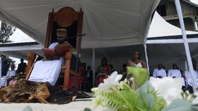 The image shows a man, wearing a white robe and a leopard-print hat, sitting on a wooden throne. He is reading from a book. The throne is covered in a leopard-print rug. The man is seated in a large white tent, surrounded by other people. There is a microphone in front of him. In the background, there is a white building and a grassy area. There is a large bouquet of white flowers in the foreground, partially obscuring the view. The scene is in Rukoko, Democratic Republic of the Congo, at coordinates -1.624, 29.258. The man may be a leader, or a figure of authority. The people gathered around him may be his supporters or subjects. The event may be a ceremony or a meeting. The white tent and flowers suggest that the event is celebratory.