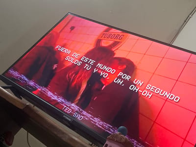 The image is a close-up, slightly angled shot of a television screen displaying a scene with text overlays. The screen shows a red-toned background with a grid pattern and what appears to be silhouettes of two people in an embrace. A prominent logo for "TUBORG" is visible in the upper right quadrant of the screen. White, pixelated text appears across the lower portion of the screen, reading "FUERA DE ESTE MUNDO POR UN SEGUNDO SOLOS TÚ Y YO, UH, OH-OH" and "TED-SAD". The bottom edge of the TV, showing the "HYUNDAI" logo, is also visible. The image is taken indoors, with the TV placed on a wooden surface. The scene does not contain any identifiable features of the city of San Antonio, Venezuela.