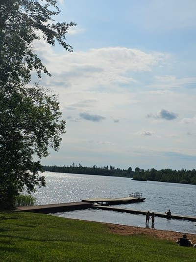 The image depicts a serene lakeside scene in Smålandsstenar, Sweden, on a bright, partly cloudy day. In the foreground, a lush green lawn slopes down to a sandy beach bordering a tranquil body of water. Several individuals are present: two children stand in the shallow water near a wooden pier, holding hands. Further along the pier, another person is seated. In the background, a dense forest lines the opposite shore, with trees extending into the distance. The water reflects the bright sky and sunlight, creating a shimmering effect. The overall atmosphere is one of peaceful recreation in a natural setting.