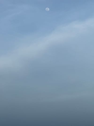 A full moon is visible in the daytime sky, set against a backdrop of light blue and grey clouds. The moon is a small, bright sphere on the left side of the frame, with subtle shading indicating its texture. Wispy clouds stretch across the sky, creating a soft, diffused light. The overall atmosphere is serene and calm. The image captures a moment of natural beauty, highlighting the presence of the moon in daylight. The location context is Arugam Bay, Sri Lanka.