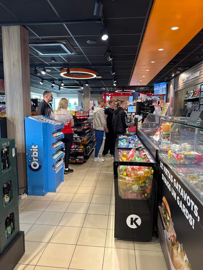 The image depicts the interior of a Circle K convenience store in Saldus, Latvia. Four individuals are standing in a line, presumably waiting to pay at the checkout counter. A blue Orbit gum display is positioned at the front of the line. Shelves stocked with various products such as snacks, drinks, and groceries line the walls. The store has a modern design with track lighting, black ceiling tiles, and an orange stripe along the upper part of the wall. A sign above the refrigerated section promotes "UZ VIETAS GATAVOTS, SVAIGS UN GARDS!", which translates to "Made on the spot, fresh and delicious!".