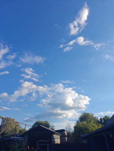 The image depicts a rural scene, likely a backyard in Kotelva, Ukraine, under a bright blue sky with scattered white clouds. In the foreground, there are several structures, including a dark-colored shed or garage with an open bay revealing items inside, and parts of another building with a corrugated roof. Trees, predominantly green deciduous ones, form a dense backdrop behind the structures, with some appearing drier and bare. A metal frame structure with horizontal bars is visible on the left side, possibly a clothesline or support. A small white vehicle, possibly a car or cart, is partially visible in the open bay of the shed. The lighting suggests it is daytime, with sunlight illuminating the clouds and casting shadows on the ground and structures. There are no people visible in the frame.