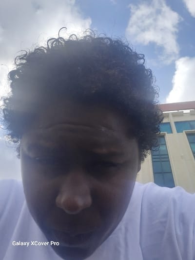 The image depicts a close-up, upward-angled view of an individual with dark skin and short, dark, curly hair, wearing a white t-shirt. The person's head is tilted forward, and facial features are partially obscured by shadows. The background shows a blue sky with scattered white clouds. To the right and behind the individual, a multi-story, light-colored building with multiple rectangular blue-tinted windows is visible. This building is identified as the Tulsa County Courthouse, located at 500 S Denver Ave W, Tulsa, Oklahoma, United States of America. A "Galaxy XCover Pro" watermark is present in the lower-left corner of the image. The scene is outdoors under daylight conditions.