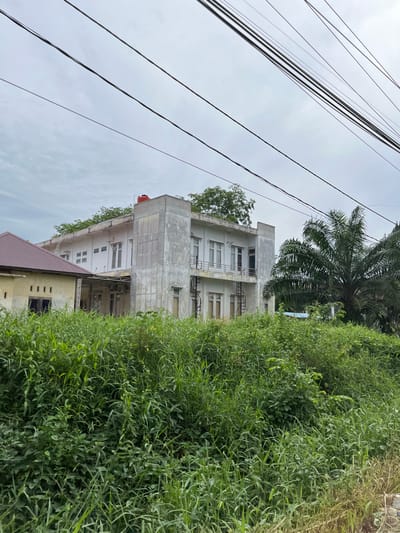 This media file captures a street-level view of a building and its surroundings, oriented vertically. The primary subjects are a multi-story building, a dense patch of green vegetation, and a cluster of overhead electrical wires against a cloudy sky.
The building appears to be a light-colored, possibly concrete structure with multiple floors. It has numerous windows, some with visible frames and others appearing as dark openings. There are also external staircases visible on one side of the building, suggesting it might be an apartment complex or an older office building. A red water tank is perched on the roof of the upper section of the building. The vegetation is lush and overgrown, dominating the right side of the frame, with some plants reaching up towards the building and encroaching on its lower levels. A palm tree is partially visible in the upper right corner, hinting at a tropical or subtropical climate.
The scene appears to be outdoors during the daytime, with an overcast sky suggesting cloudy weather. The electrical wires are a prominent feature, crossing diagonally across the upper portion of the image, indicating proximity to a residential or commercial area with active utilities. There is no visible text in the image. The overall impression is one of a slightly neglected or overgrown urban or semi-urban environment in Bundar, Indonesia, with a focus on the juxtaposition of built structures and natural growth. No people or specific activities are depicted.