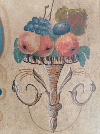 The image is a close-up of a faded mural painting on a textured wall, possibly a ceiling or wall in a building. The mural depicts a still life arrangement of fruit in an ornate vase. At the top of the vase, there are bunches of dark blue grapes and several red and orange fruits, likely apples or peaches, with visible dark pits. To the right of the fruit, there is a patch of yellow and reddish-brown foliage or perhaps another cluster of fruit, also faded. The vase itself is depicted in shades of brown and white, with a striped rim. The lower part of the mural features decorative scrollwork in brown, white, and black, extending outwards from the base of the vase. The overall impression is one of age and wear, with the paint peeling in several areas, revealing the textured surface beneath. The background is a neutral, beige color. There are no people or animals present in the image. The painting appears to be a decorative element within a room, and its style suggests it could be part of historical or traditional decor. The location in the city of Grünwald, Germany is not visually discernible from the image itself, and would require external information to determine.