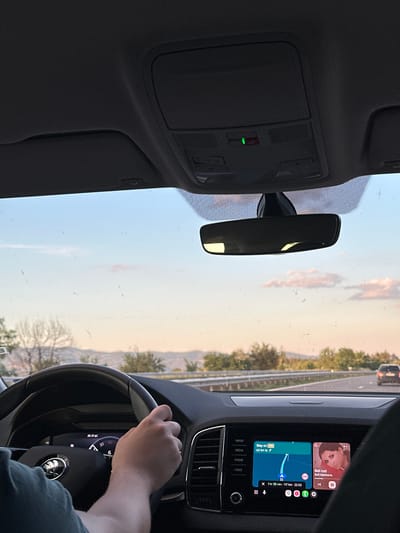 The image shows the interior of a car driving on a highway. A person's hands are on the steering wheel, and the car's dashboard displays a navigation system and music player. The navigation shows a route with 60 km remaining to the destination, indicating a travel time of 1 hour and 25 minutes. The music player displays the song "Bell rozi" by Sofi Marinova. Outside the car, the scenery suggests a rural or mountainous landscape with trees and hills under a sky with scattered clouds, typical of the region around Gorno Varshilo, Bulgaria, especially during the golden hour of sunset. There are also other cars visible on the highway in the distance. The car's interior features a rearview mirror and the car's overhead console with an illuminated green light, possibly an indicator for a safety system. The overall scene depicts a journey in progress during the late afternoon or early evening.