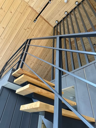 The image shows an angled view of a modern architectural structure, likely an outdoor patio or deck, with a focus on the interplay of wood and metal. The primary subjects are the wooden ceiling planks and the dark gray metal railings and beams. The wooden ceiling is constructed from narrow planks with visible grain and knots, giving it a natural and rustic feel. Overlapping this is a geometric arrangement of dark gray metal elements, including thin vertical bars forming a railing or screen, and thicker horizontal beams. These metal components create a sense of depth and modernity.

Supporting the metal structure are several thick, light-colored wooden beams, positioned vertically and angled, with visible wood grain and texture. These beams are attached to the metal framework. The background shows a section of dark gray siding, suggesting the exterior of a building.

There are several circular light fixtures embedded in the wooden ceiling, some of which are illuminated, suggesting it might be evening or a dimly lit area. The lighting casts subtle shadows on the wooden surfaces.

No people or specific activities are depicted in the image. The scene appears to be static, focusing on the structural and material elements of the architecture. The location is implied to be Novaya Vozhoika, Russia, but this context does not directly inform the visual description of the image itself. There is no visible text in the image. The overall impression is one of clean lines, a blend of natural and industrial materials, and a contemporary design aesthetic.
