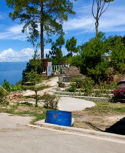 The image depicts an outdoor scene in Parik Sabungan, Indonesia, featuring a terraced landscape with a large tree in the foreground. The terrain is covered with sparse grass, dirt, and some gravel. In the mid-ground, a winding concrete path leads up to a slightly elevated area with stone retaining walls and steps. Several bushes and small trees are planted along the path and terraces. A prominent blue cylindrical object, resembling a large barrel, sits on a concrete base near the bottom of the image. In the background, a body of water, likely a lake, is visible, bordered by distant hills. The sky is blue with scattered white clouds. The overall impression is of a scenic overlook or park area.