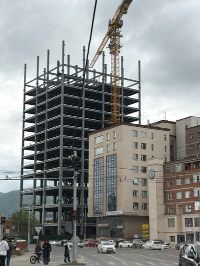 In Ulaanbaatar, Mongolia, a multistory building under construction with a steel frame dominates the center of the image. A yellow construction crane is positioned to the right of the building. Adjacent to the construction site, a modern tan building with multiple windows is visible. Across the street, a traffic intersection with a pedestrian crossing is present, with several vehicles on the road. Pedestrians are crossing the street or walking on the sidewalk. The sky is overcast with clouds.
