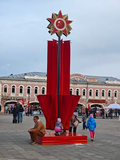 A wide shot shows a public square in Uglich, Russia, with a large red star and banners memorializing the Soviet Union. In the foreground is a red platform, next to which a man sits and a child kneels. Further away, a woman and child walk, with other people and street vendors visible in the background. A light pink building with many windows is behind the people in the square, with the word "HOTEL" and other Cyrillic text on top of it. The sky is overcast and gray.