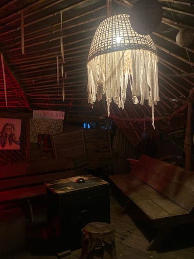 The image depicts the interior of a building in Baan Don Sai, Thailand. The structure appears to be constructed primarily of wood and bamboo. A unique chandelier-like light fixture hangs from the ceiling, with a woven shade and tattered white fabric strips.
To the right, a wooden bench or table is visible along with a dark box-like object. Further back, on the wall, are a sign with the words "Drink with me, darling", a patterned wallpaper, and framed images. The environment is dimly lit with a rustic, possibly bohemian, aesthetic.