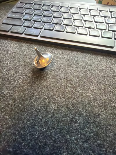 The image displays a close-up, angled view of a workspace, likely a desk, featuring a dark-colored keyboard and a metallic spinning top resting on a textured, grey surface.

The primary subject on the left is a portion of a keyboard, oriented vertically. It has dark grey keys with white lettering. The keyboard layout is QWERTY with secondary Hebrew characters, such as 'ש' (Shin), 'ד' (Dalet), 'ג' (Gimel), 'כ' (Kaf), 'ע' (Ayin), 'ח' (Chet), 'י' (Yod), 'ל' (Lamed), 'ם' (Mem Sofit), 'פ' (Pei), 'ף' (Pei Sofit), 'ת' (Tav), 'צ' (Tsadi), 'מ' (Mem), 'נ' (Nun), 'ב' (Bet), 'ס' (Samekh), 'ז' (Zayin), 'ק' (Kof), 'ר' (Resh), 'א' (Aleph), 'ט' (Tet), 'ו' (Vav), 'ן' (Nun Sofit). Visible English function keys include "tab," "caps lock," "shift," "ctrl," "start," "opt," "alt," and "cmd".

To the right of the keyboard, positioned centrally in the frame, is a small, silver-toned metallic spinning top, or a dreidel-like object. It has a shiny, reflective surface, indicating a polished metal construction, and is resting on its side with its pointed stem facing upwards and slightly left.

The surface beneath the keyboard and spinning top is a dark grey, highly textured material resembling a dense, short-pile carpet or a fabric desk mat. The lighting appears to be even, likely indoor, casting soft reflections on the metallic top. The presence of Hebrew characters on the keyboard strongly suggests the location is in Israel, aligning with the provided context of Kafr Yasif.