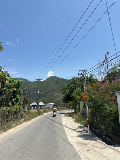 The image depicts a street in Diên Khánh, Vietnam, featuring a two-lane asphalt road curving to the right. A motor scooter with two people is traveling along the road. The road is bordered on the right by a concrete wall and vegetation, and on the left by trees and utility poles with power lines extending across the blue sky. A mountain is visible in the background. A road sign is present.
