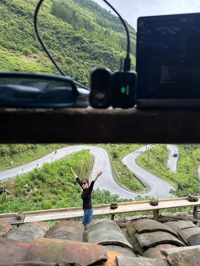 The image depicts a woman standing on what appears to be a rooftop or platform made of wooden planks and clay tiles. She is wearing a black long-sleeved shirt, blue jeans, and sunglasses. Her arms are outstretched.

Below the platform, a winding road cuts through a green, hilly landscape. A vehicle is visible on the road.

In the foreground, inside what seems to be a room or interior space, there's a laptop computer, two small black devices connected by a cable, and part of a microphone.

The location is Yên Minh Commune, Vietnam, evidenced by the topography and distinctive curving road.