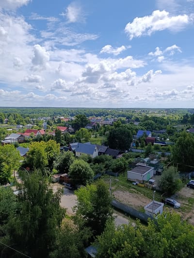 The image captures an aerial view of a rural village or settlement surrounded by dense green forests under a bright, partly cloudy sky. The scene is set in Alexandrov, Russia. The time of day appears to be late morning or early afternoon, indicated by the strong daylight and clear shadows. The weather is pleasant, with a mix of sun and scattered white clouds in a blue sky, suggesting a warm summer day.
The foreground is dominated by lush green trees, some with lighter green foliage and others with darker, denser canopies. Below the trees, a scattering of houses with various roof colors, including red, blue, and grey, are visible. These homes appear to be single-family dwellings, many with fenced yards. Several of the houses have visible windows and some have chimneys.
Further into the scene, the village expands with more houses and buildings interspersed with trees and open grassy areas. Roads and pathways wind through the settlement. In the middle ground, a few vehicles, including a dark-colored car, are parked on a dirt or gravel area. There are also some outbuildings and what appear to be small greenhouses or garden structures. The overall impression is a tranquil, residential area nestled within a natural landscape. No people are clearly visible in the image. There is no discernible text in the image.