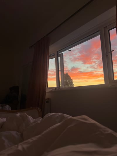 The image depicts a bedroom interior at dusk, with a view of a colorful sunset through an open window. The foreground is dominated by a rumpled duvet and pillows, suggesting a person is in bed. To the left, a patterned curtain is drawn partly closed. Beyond the window, a vibrant sky displays hues of orange, pink, and yellow clouds, with the silhouette of a tree visible. A radiator is visible beneath the window. The room appears to be dimly lit by the fading sunlight. The specific location in Brighton, United Kingdom, cannot be determined from the visual information provided.