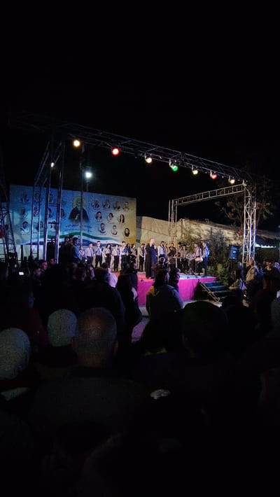 A nighttime scene shows a crowd watching a performance on a stage. The stage is illuminated by overhead lights and a backdrop with images of people and Arabic text. Performers in uniform are on the stage along with a person in black robes. The audience, mostly silhouettes due to the darkness, are focused on the event. A metal truss structure supports the lighting. The lighting includes colors such as green, yellow and red.