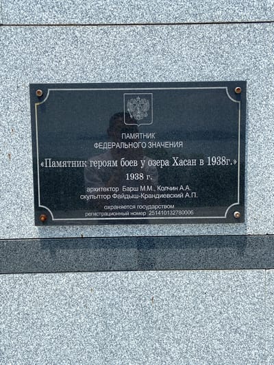 This is a close-up, eye-level, outdoor, daylight, slightly overcast photograph of a black granite memorial plaque mounted on a grey speckled granite wall. The plaque is rectangular with decorative curved corners and is secured by four visible screws, one at each corner. The text on the plaque is in white, engraved lettering.

The inscription reads:

ПАМЯТНИК
ФЕДЕРАЛЬНОГО ЗНАЧЕНИЯ

«Памятник героям боев у озера Хасан в 1938г.»

1938 г.
архитектор Барш М.М., Колчин А.А.
скульптор Фаидыш-Крандиевский А.П.

охраняется государством
регистрационный номер 251410132780006

At the top of the plaque, the Russian coat of arms is embossed. A partial reflection of a person is visible on the surface of the plaque, obscuring some of the inscription. The background wall is made of large granite slabs with a visible horizontal seam near the top of the plaque. The environment appears to be a public space, likely a memorial site. The overall scene is static and formal, typical of a historical monument. The location is confirmed as Kraskino, Russia based on contextual information not visible in the image itself.