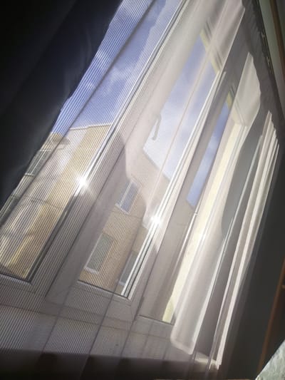 The image shows a view through a window with sheer curtains. The curtains are white and translucent, with vertical pleats. Sunlight streams through the window, creating bright highlights and shadows. Outside the window, there is a building with a yellow facade and several windows. The sky is blue. The perspective suggests the photo was taken from inside a room looking outwards. The location is specified as Geranionski selski Soviet, Belarus. There are no people or animals visible in the image. The scene appears to be a domestic or residential setting, possibly an apartment or house.