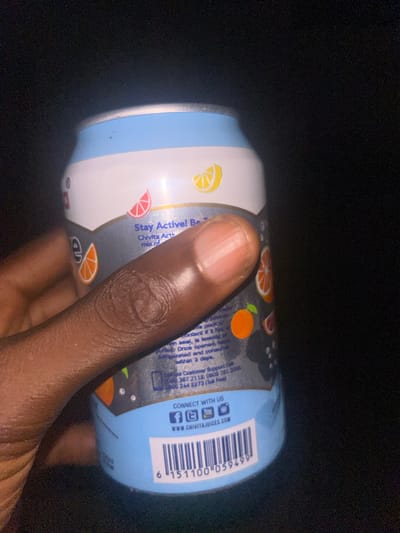 In Egbe, Nigeria, a close-up image depicts a dark-skinned hand holding a Chivita Active beverage can. The can features a light blue and white top and bottom, with a dark grey mid-section displaying illustrations of sliced lemons, grapefruits, and oranges. Prominently visible text on the label includes "Stay Active!", "Chivita Active", customer support telephone numbers (0806 387 2113, 0809 761 2000, 0800 244 2273), social media icons with "CONNECT WITH US", and the website "WWW.CHIVITAJUICES.COM". A UPC-A barcode with the number 6 151100 059499 is visible at the bottom. The individual's index finger wraps across the can's label from the right, with the thumb partially visible on the left side. The background is uniformly dark, indicating low light conditions.
