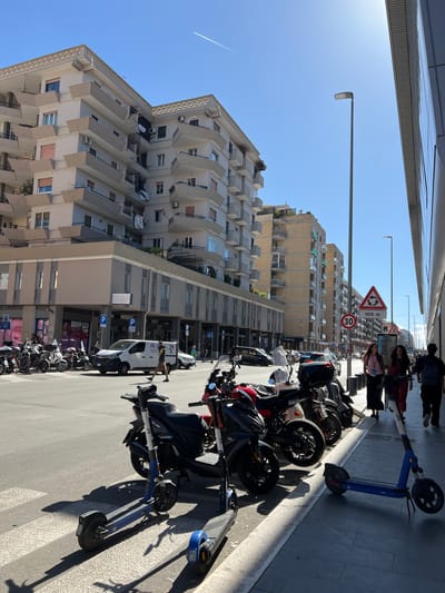 This image captures a bright, sunny street scene in a city, likely Bari, Italy, given the architectural style and urban activity. The perspective is looking down a street lined with tall, multi-story buildings. On the left, a light-colored residential or mixed-use building dominates the frame, characterized by numerous balconies and visible air conditioning units. Further down the street, another building exhibits a more complex, modular facade.
The street is a bustling thoroughfare, with a significant number of vehicles parked along the right side. A long row of motorcycles and scooters, many in shades of black, red, and silver, fills the parking spaces. Several blue and white shared electric scooters, some bearing the brand name "BIT," are also visible on the sidewalk and street. A white commercial van, with "db Service" or "db S.r.l." text and a logo, is parked further away.
Several people are present on the wide sidewalk on the right, some walking or standing near the parked vehicles. The bright, clear blue sky and the long, sharp shadows cast by the buildings and vehicles suggest it is either mid-morning or afternoon.
Notable details include European road signs: a triangular sign indicating a roundabout ahead, with "100 m" printed below it, and a circular red-bordered sign indicating a "30" km/h speed limit. Blue square 'P' parking signs are also visible. On a grey building, a sign displays "YVES SEGAL" or "YVES SEGALA" and another partially visible sign below it begins with "I PUNTI DI RIFERIMEN". A faint airplane contrail is visible in the clear sky. The overall scene depicts a typical, busy urban environment.