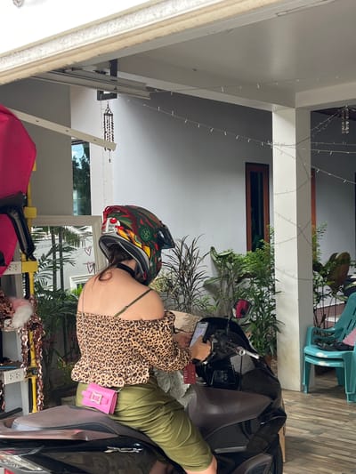 The image shows a woman wearing a leopard print top, olive green pants, and a colorful helmet sitting on a black motorcycle. She is holding a phone and appears to be looking at it. The motorcycle is parked under a covered patio area with a white ceiling and supports. In the background, there are plants, windows, and a teal chair stacked on another. The scene is likely in the exterior of a residence, possibly in the city of Ban Kanok 23, Thailand, given the presence of tropical plants and the style of architecture.
