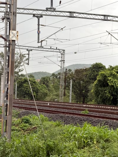 The image shows a view of a railway line in Saphale, India, with overhead power lines and supporting structures. In the foreground, a tall metal pole with a sign is visible on the left. The sign has text that appears to say "POWER LINE" and "WORK BLOCK." Several sets of parallel railway tracks run horizontally across the mid-ground. The tracks are made of dark gravel and red-brown sleepers. Beyond the tracks, dense green foliage and trees form a natural backdrop, with hills visible in the distance under an overcast sky. The scene is devoid of people.