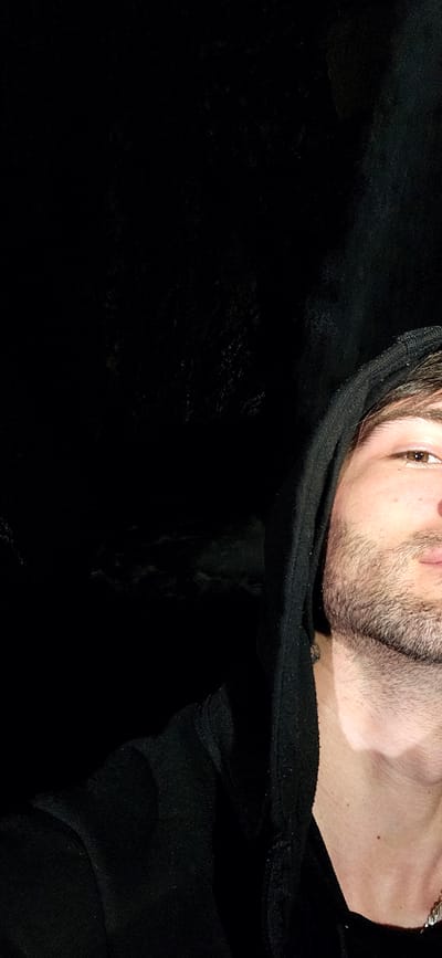 The image shows a close-up, chest-up view of a young man wearing a black hooded sweatshirt. Only the right side of his face is visible, revealing his eye, nose, and mouth. He has short brown hair and a light beard. The background is predominantly dark and out of focus, with some texture suggesting it might be a natural environment or a dimly lit indoor space. The man is looking towards the camera. The scene is not set in the city of Kostenets, Bulgaria; this image has no discernible geographic context.