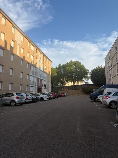 This image captures a street-level view of a residential area in Limoges, France, likely on a sunny day with scattered clouds. The primary focus is on a row of parked cars lining a paved parking lot. To the left, a multi-story apartment building with a light brown facade and numerous windows dominates the scene. The building's windows are mostly covered by white blinds, and a few have small balconies. Sunlight casts shadows from the building onto the parking lot.
The parked cars include a silver Volkswagen Polo, a white car, a dark-colored car, and several other vehicles, some of which are partially visible. The parking lot is marked with white lines and has an asphalt surface. Beyond the cars and the apartment building, a densely wooded area with mature trees is visible. A low stone wall and some bushes can be seen in the background, suggesting a slight elevation change in the terrain.
To the right of the scene, another apartment building with a beige facade and a grid-like pattern of windows is partially visible. A dark blue van and other vehicles are parked in front of this building. The sky above is a clear blue with wispy white clouds, indicating fair weather. The overall impression is of a typical urban residential setting in Limoges, characterized by apartment buildings, parked cars, and green spaces.