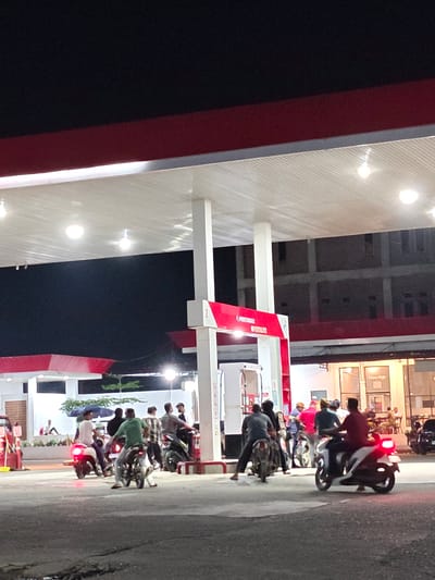 At night, a group of people are gathered around several motorcycles at a gas station in Blangmetimu, Indonesia. The gas station has a red and white canopy with fluorescent lighting. Several individuals are seated on motorcycles, with some appearing to be fueling up or preparing to depart. In the background, a building with multiple stories is visible, as are some shops. The scene is illuminated by artificial light, creating a somewhat blurry and grainy image.