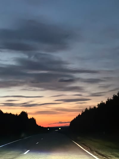 A long, empty asphalt road stretches forward, flanked by dark silhouettes of dense coniferous trees on either side. The sky above is a gradient of twilight colors, transitioning from deep blues and grays with scattered clouds at the top to a vibrant band of orange and yellow just above the horizon. The road has visible white lane markings, and the headlights of the vehicle from which the photo is taken illuminate the immediate foreground. The scene appears to be a rural landscape, possibly on the outskirts of Крынды, Russia, during dusk. There are no people or animals visible in the image.