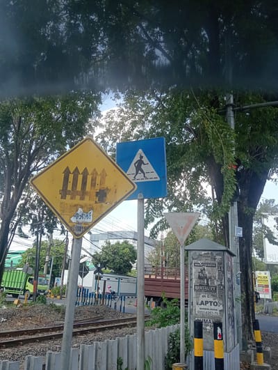 The image depicts a street scene in Banjarkemantren, Indonesia, focusing on a railway crossing. In the foreground, railway tracks are visible, bordered by a concrete barrier and gravel. A grey pole with black and yellow striped bases stands prominently in the center of the image.

Several traffic signs are mounted on poles. A yellow diamond-shaped sign with a black symbol of a fence and three arrows pointing upwards is on the left. To its right, a blue square sign with a white triangle containing a black silhouette of a pedestrian walking on a crosswalk is displayed. Below the pedestrian sign is a white triangular sign with a red border and a red inverted triangle symbol.

A utility box, plastered with various advertisements and flyers, is situated to the right of the signs. One flyer advertises "MAN OF THE MATCH," another "BRUTALITY," and another a service with phone numbers. Another sign on a pole to the far right reads "POLSEK BUDURAN."

In the background, a green truck is seen turning, and a brown flatbed truck is parked near the railway crossing. A small white building with blue and white striped barriers is also visible at the crossing. The sky is clear blue, with the canopy of trees overhead casting shadows. The overall scene suggests an active transportation area with both road and rail infrastructure.