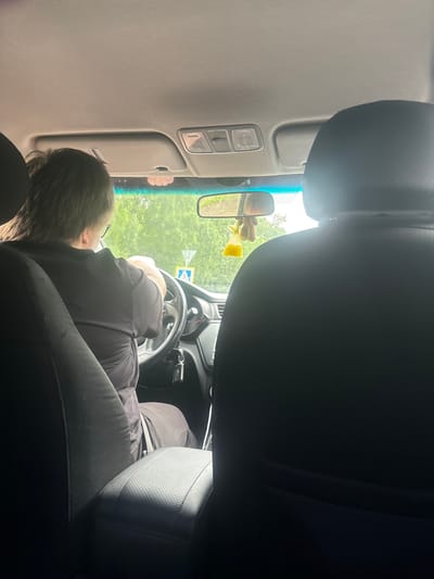 An interior view from the backseat of a car shows a person driving, facing forward with their hands on the steering wheel. They are wearing a black t-shirt. Outside the car, trees and a street sign are visible through the windshield. A yellow air freshener hangs from the rearview mirror. The car appears to be in motion or parked on a street in Kirov, Russia.